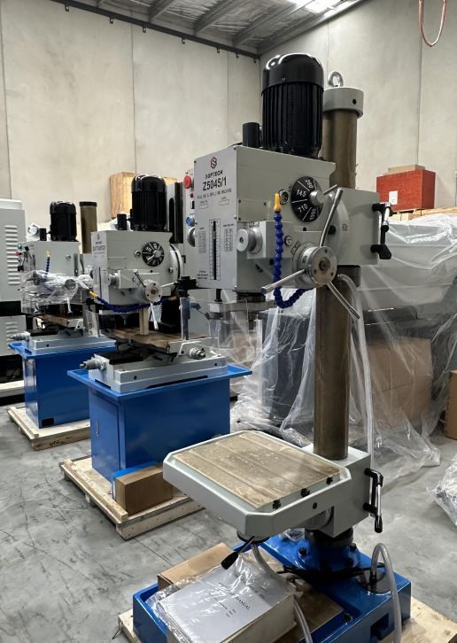 Row of industrial drill presses in a workshop, with blue bases and a concrete floor under overhead lighting.