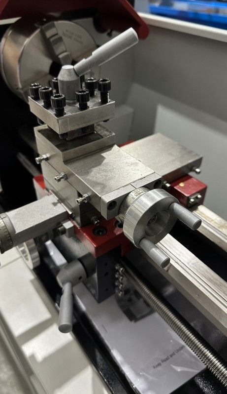 Metal lathe cutting a rectangular metal block, with toolpost and chuck visible in a workshop.
