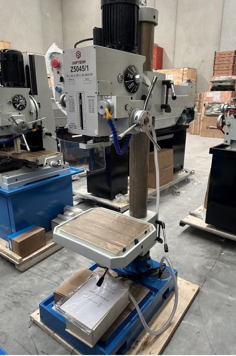Drill press machine in a workshop, with a metal worktable and blue base on a concrete floor.