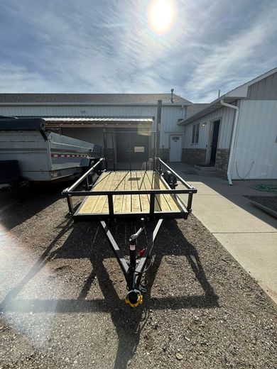 Small utility trailer in a gravel driveway beside white buildings under bright sun.