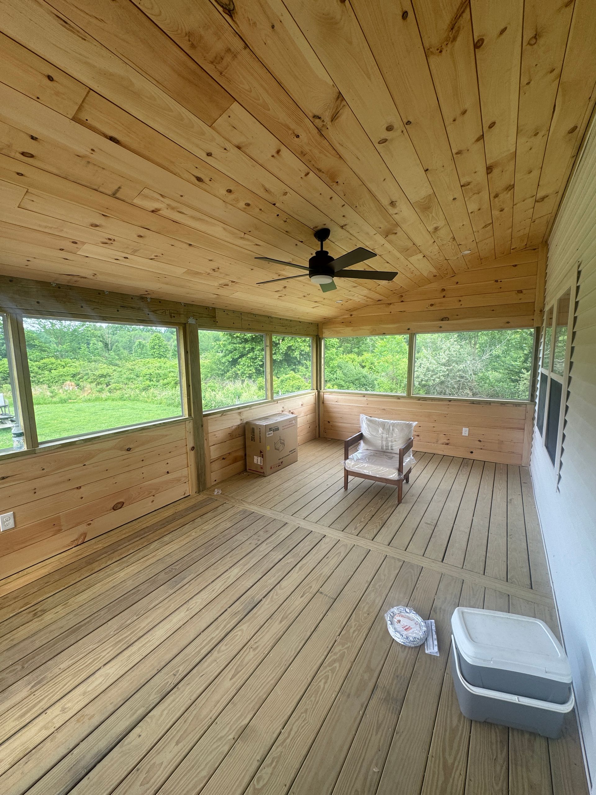 Empty wooden screened porch with ceiling fan, chairs, and green wooded view