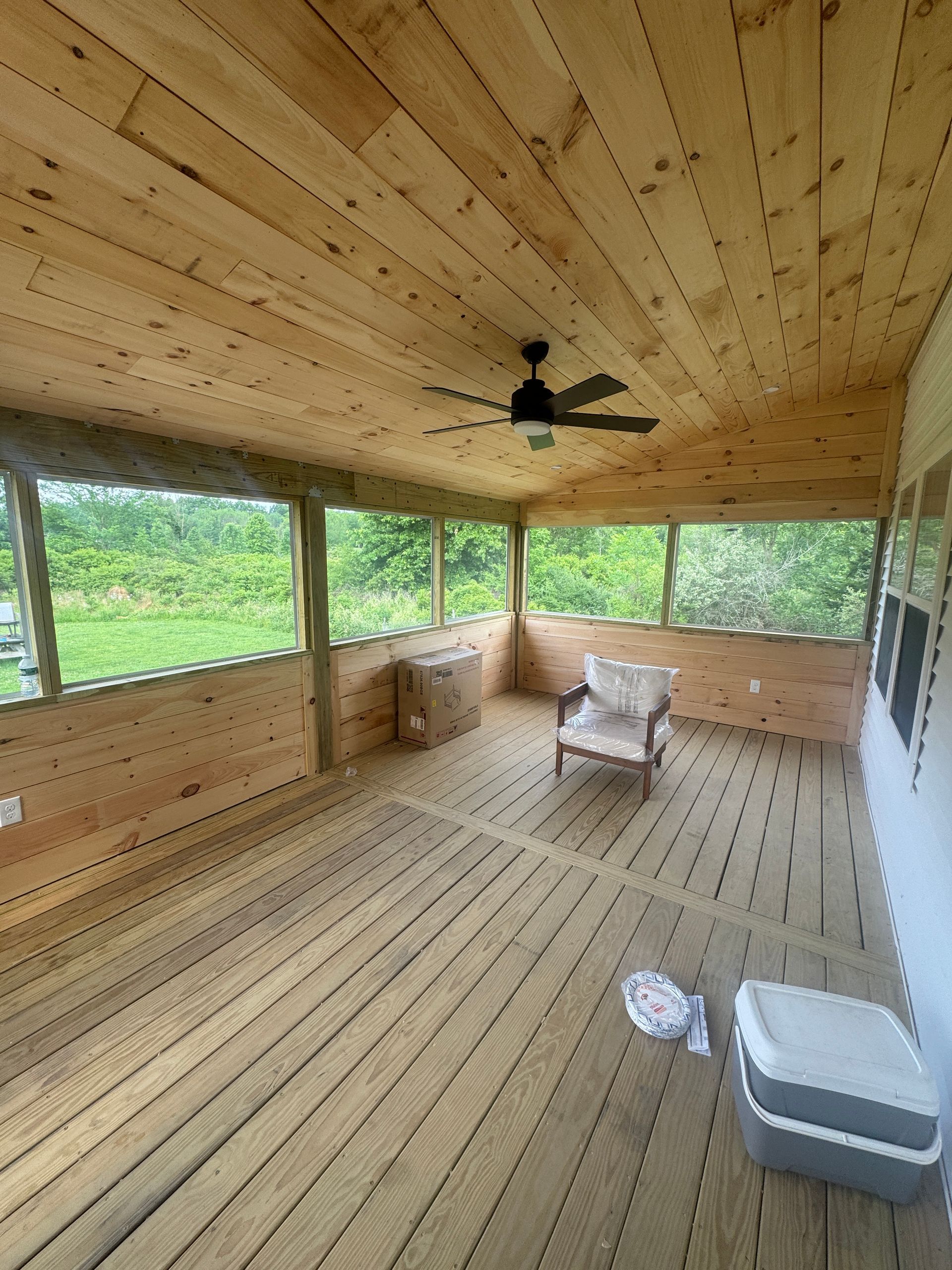 Wooden screened porch with ceiling fan, chairs, and cooler overlooking green trees