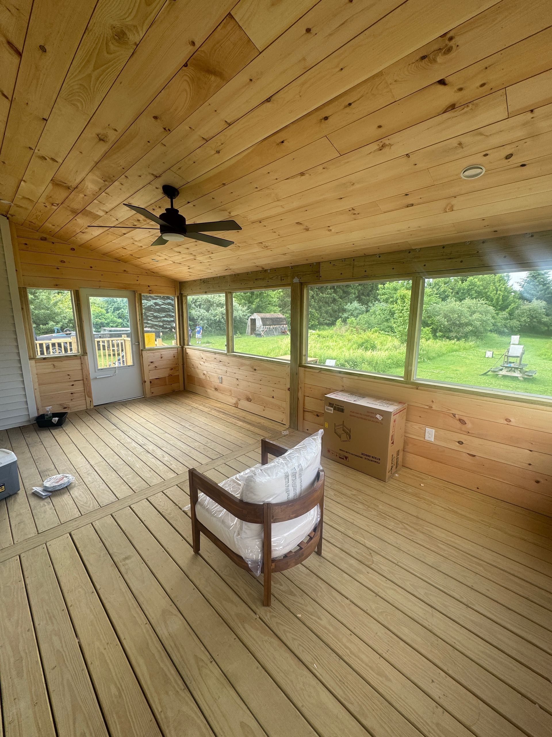 Wood-paneled screened porch with ceiling fan, chair, and windows overlooking a green yard