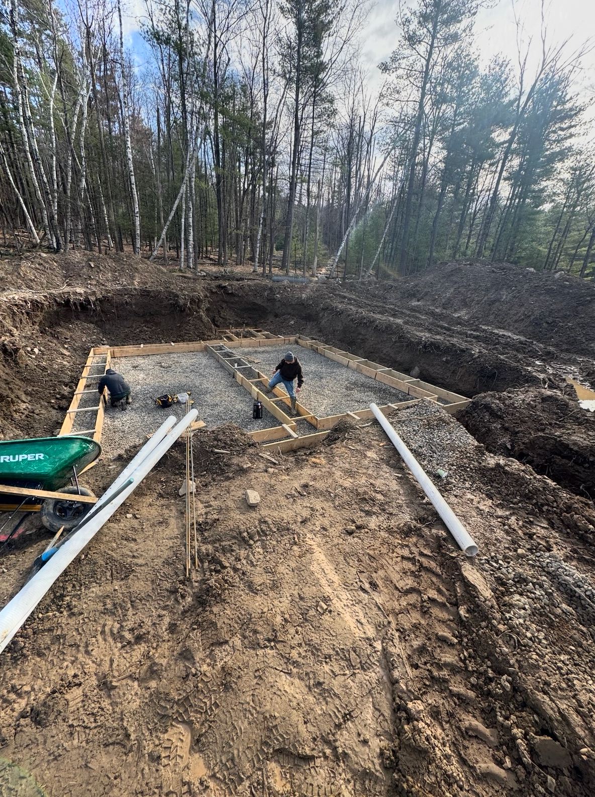 Construction site with excavated trench, gravel base, and white PVC pipes laid in the ground.