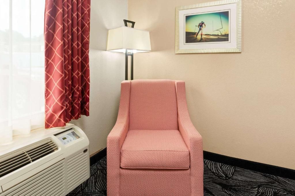 Cozy hotel room corner: pink armchair, lamp, window with red curtain, framed artwork, and heating unit.