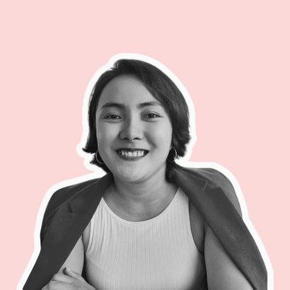 A smiling person with a blazer draped over their shoulders against a light pink background.
