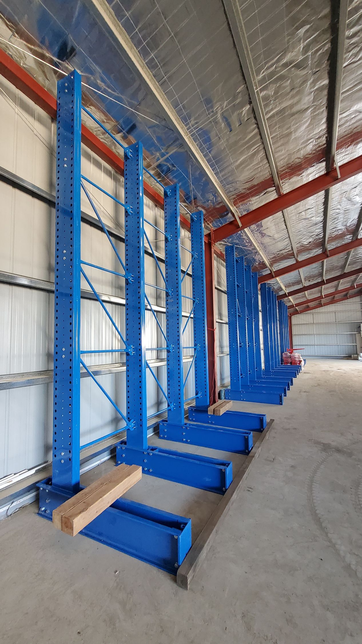 Blue warehouse rack frames lined up in an industrial building with a concrete floor and metal roof