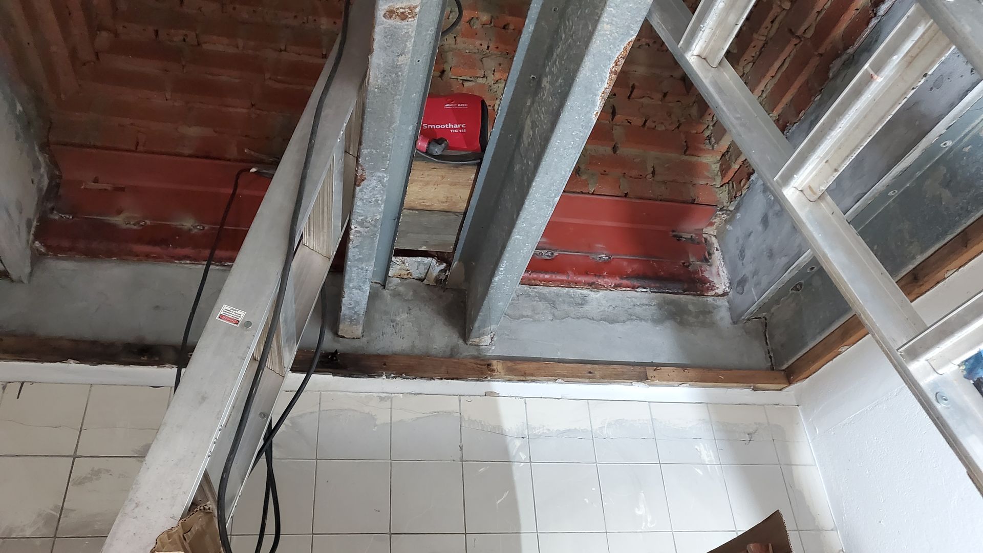 Attic view with exposed beams, red roof panels, and a small red device on the wall above white tiles