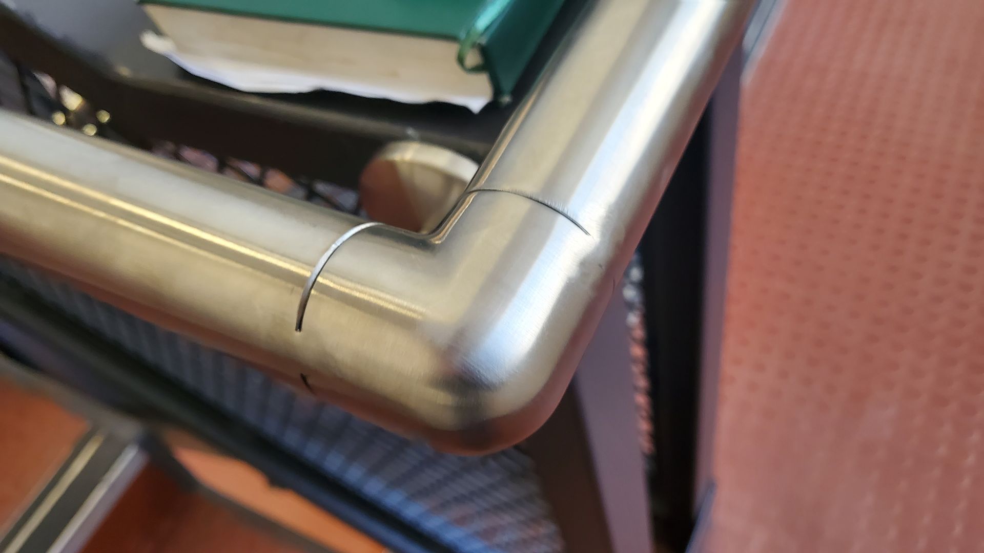 Close-up of a silver metal chair arm with a small crack, beside a green seat cushion and red flooring.