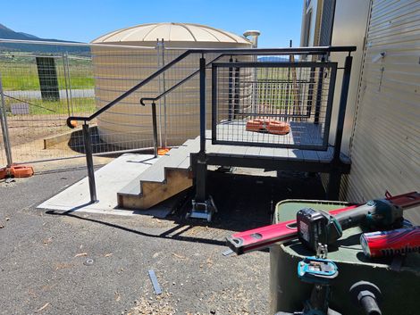 Outdoor metal stairs and black platform next to a beige tank, with tools on gravel ground.
