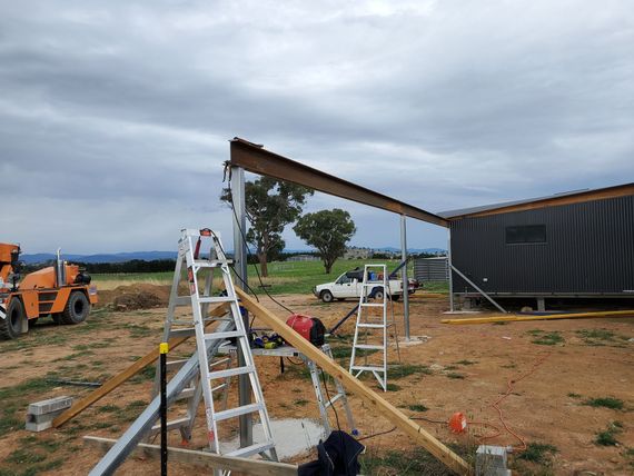 Workers use ladders and wooden beams to build a roof on a rural structure under cloudy skies.