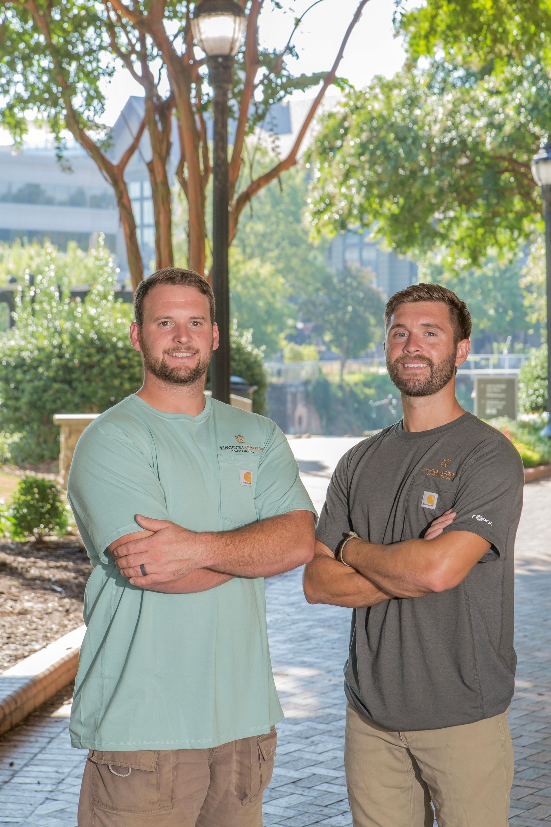 Two men with arms crossed stand on a tree-lined campus walkway in daylight.
