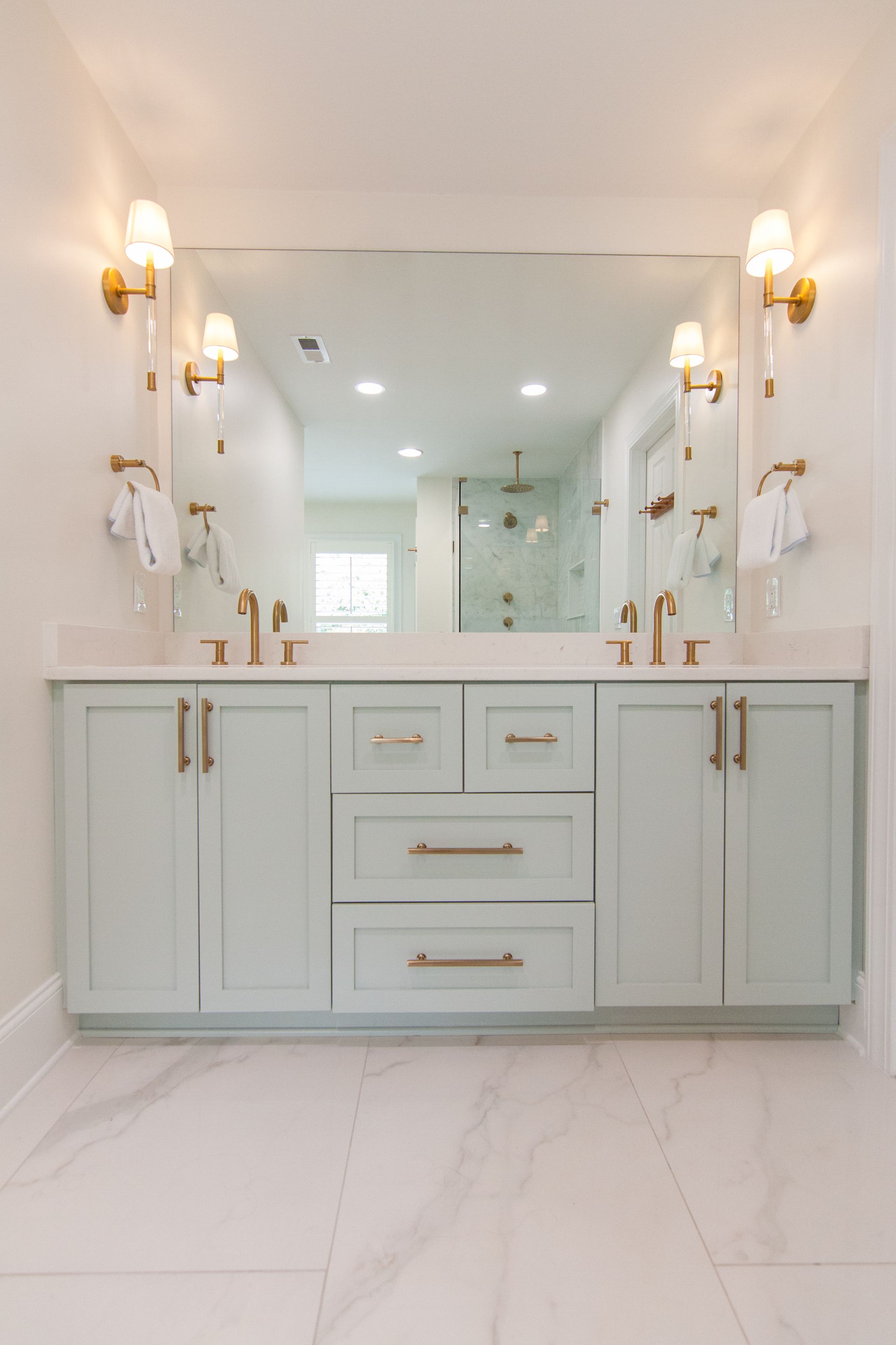 Bright white bathroom vanity with double sinks, gold sconces, and marble-style flooring and walls