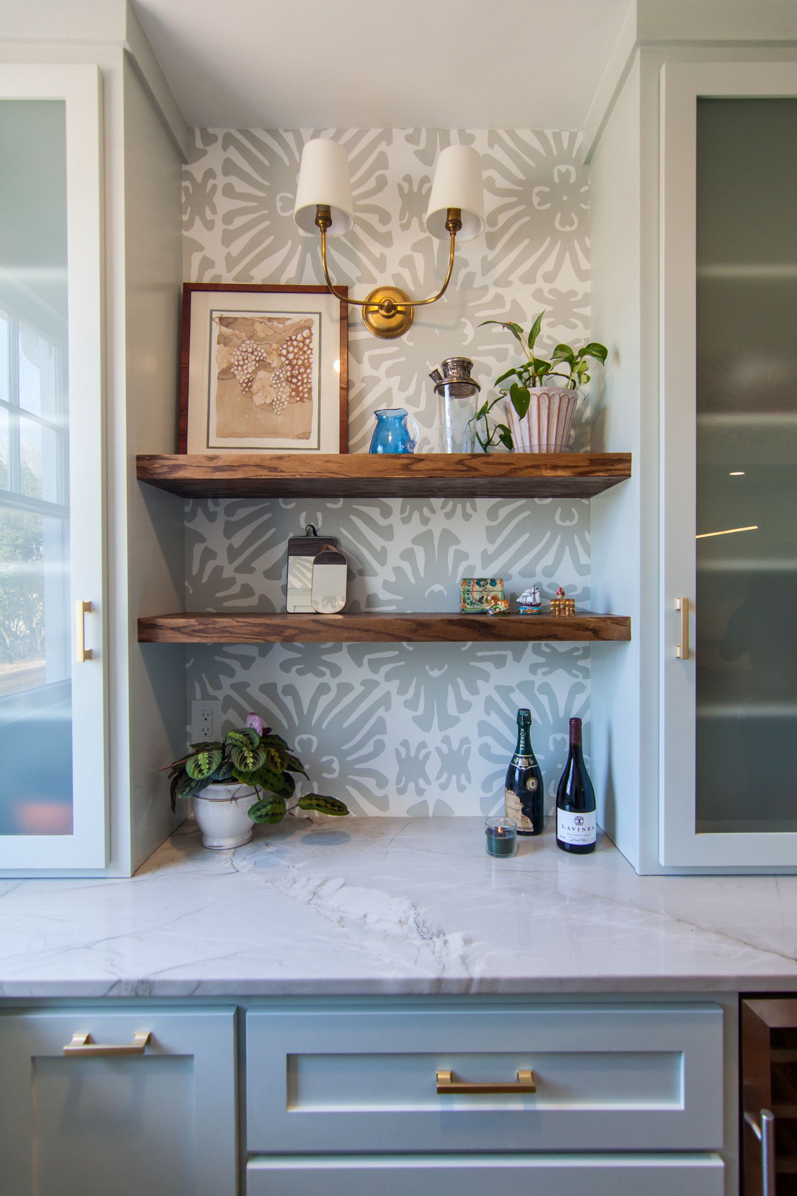 Kitchen countertop with floating shelves, plants, framed art, and decorative bottles in a light gray interior