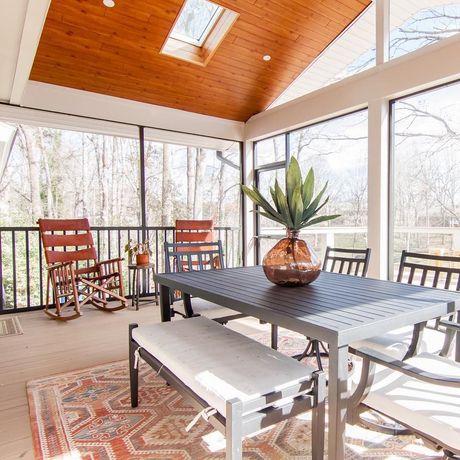 Covered porch with black dining table, bench, rocking chairs, and potted plant overlooking snowy trees