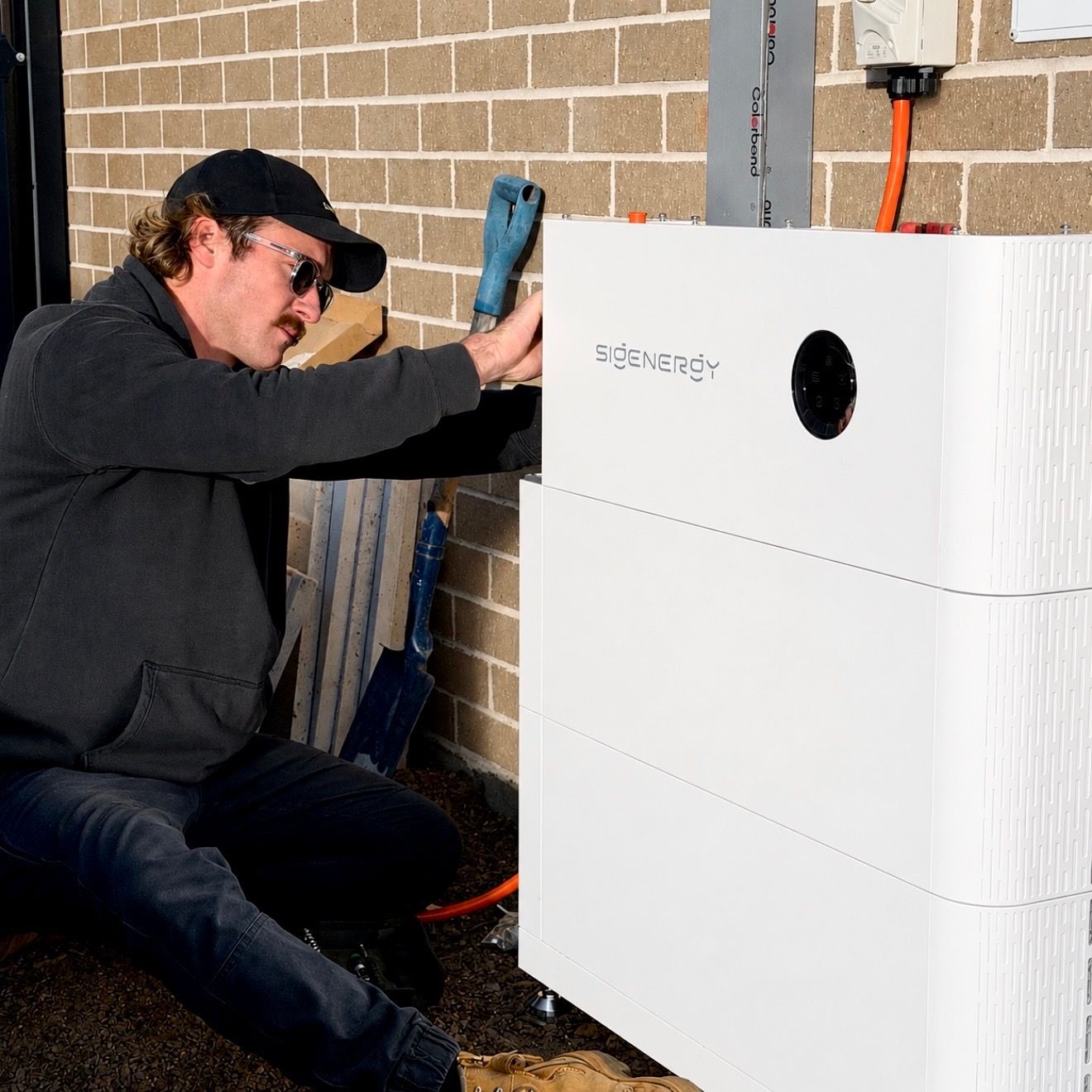 Person kneeling beside a white solar battery unit, adjusting it with a tool outdoors by a brick wall