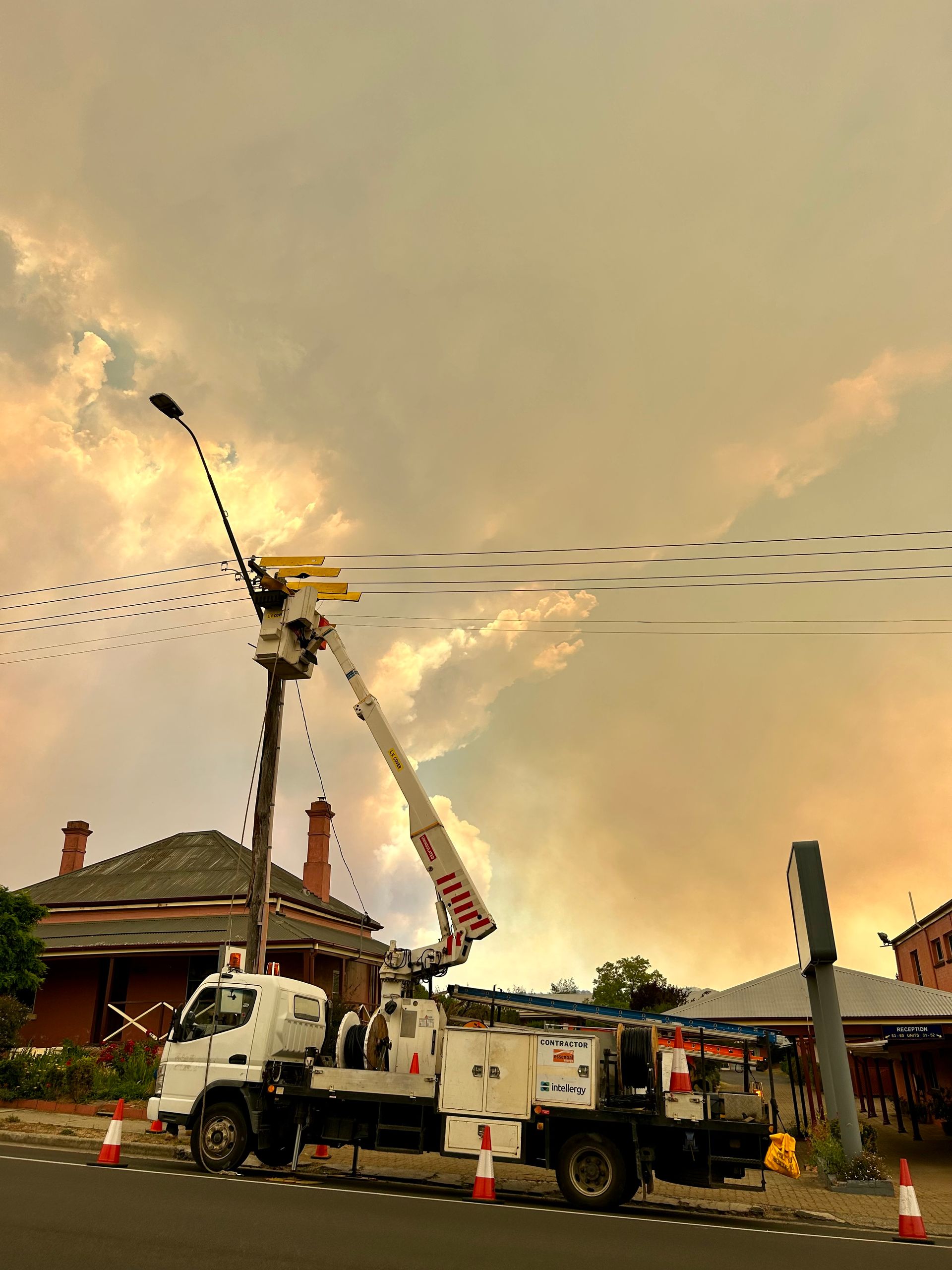 Utility trucks lift a worker on a boom near a house at sunset, with birds overhead.