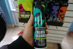 Une personne tenant une bouteille de nettoyant pour vitres Goodfellas noire et bleu sarcelle, avec d'autres produits de nettoyage pour voiture sur une étagère en arrière-plan.