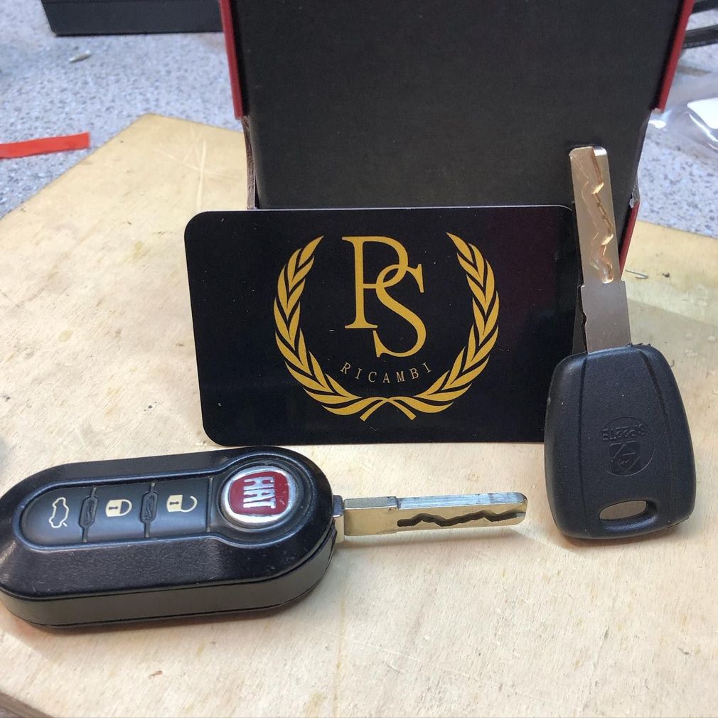 Un portachiavi a scatto nero della Fiat e una chiave di riserva posizionati davanti a un cartoncino nero con il logo PS Ricambi.