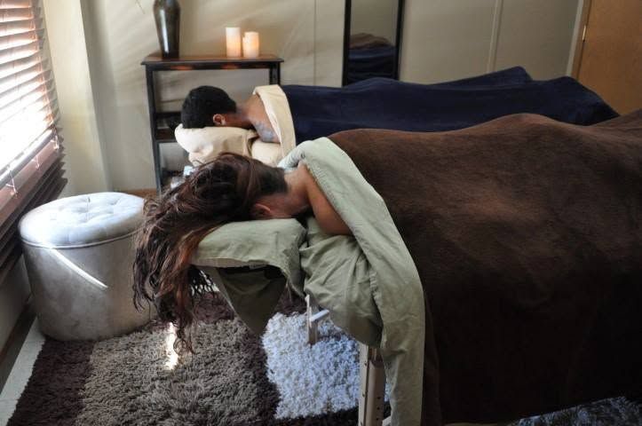 Person lying face down on a massage table in a softly lit room, covered with a blanket.