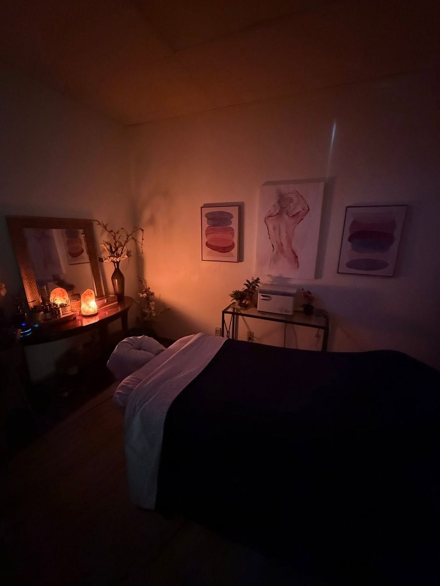 Dim bedroom with a lit candle altar, framed art, and a bed draped in dark bedding and white blanket.