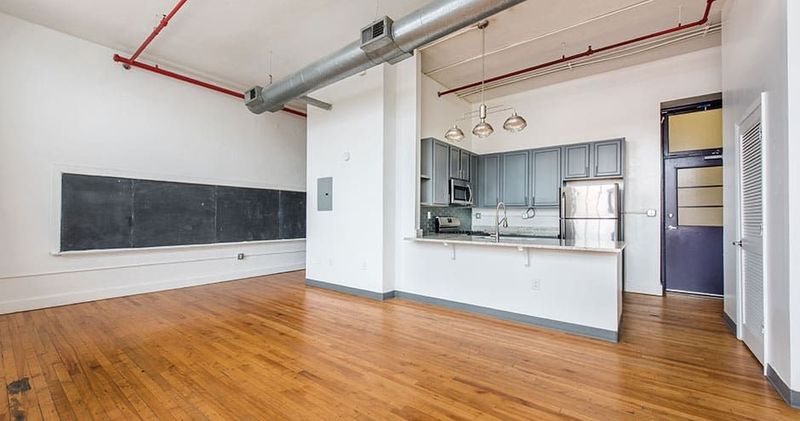 Open-concept loft with hardwood floors, kitchen, and large chalkboard wall. Exposed pipes and high ceilings.