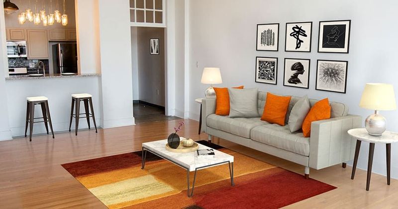 Living room with a tan sofa, orange pillows, art, and a colorful rug.