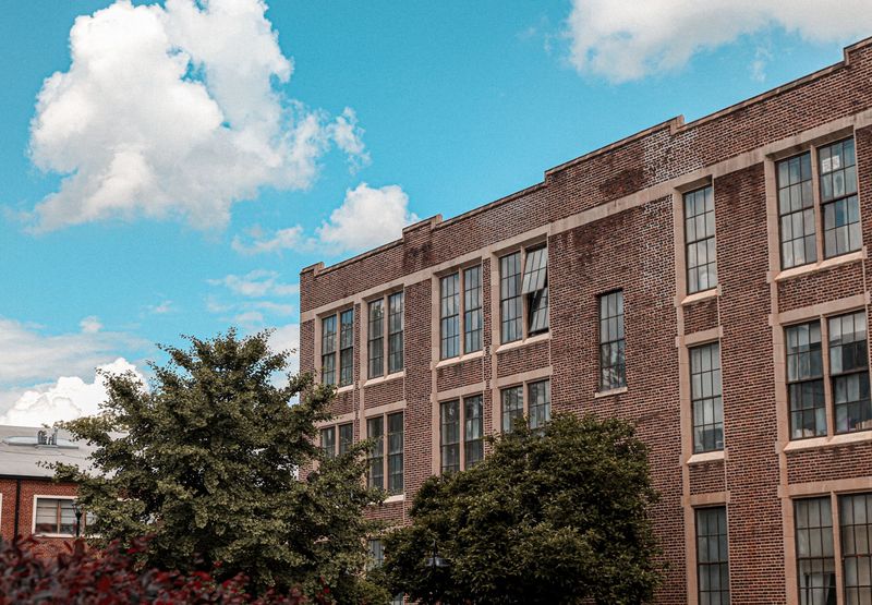 Brick building with multiple windows under a blue sky with clouds. Green trees in the foreground.