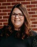 A woman wearing glasses is smiling in front of a brick wall.