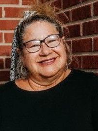 A woman wearing glasses and a black shirt is smiling in front of a brick wall.