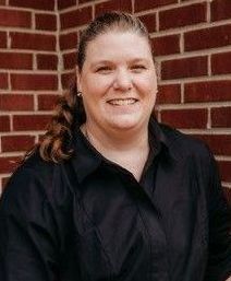 A woman in a black shirt is smiling in front of a brick wall.