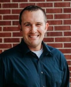 A man in a blue shirt is smiling in front of a brick wall.