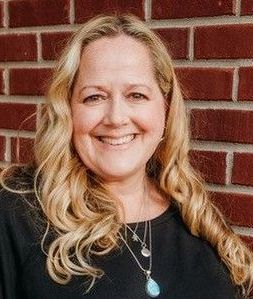 A woman with long blonde hair is smiling in front of a brick wall.