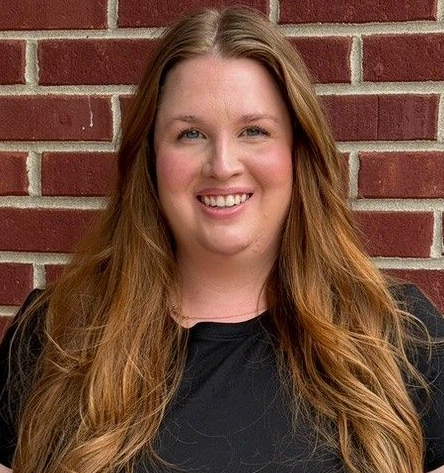 A woman in a black shirt is smiling in front of a brick wall.