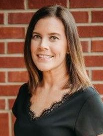 A woman in a black shirt is smiling in front of a brick wall.