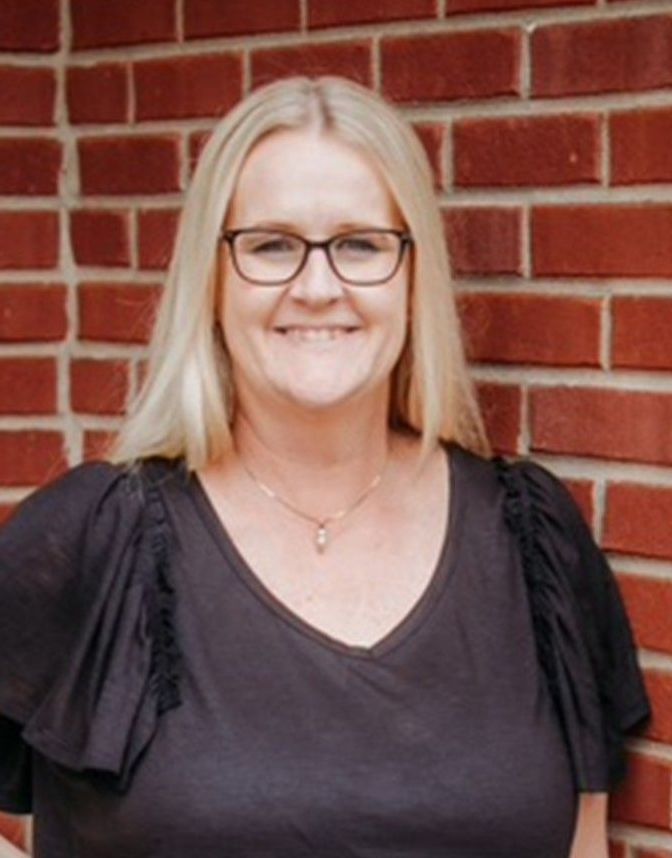 A woman wearing glasses and a black shirt is standing in front of a brick wall.