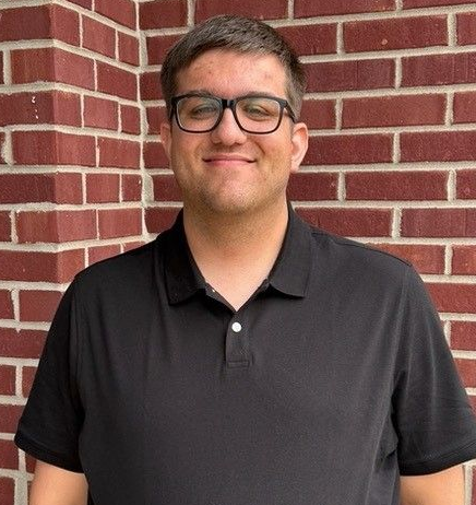 A woman wearing glasses and a black shirt is smiling in front of a brick wall.