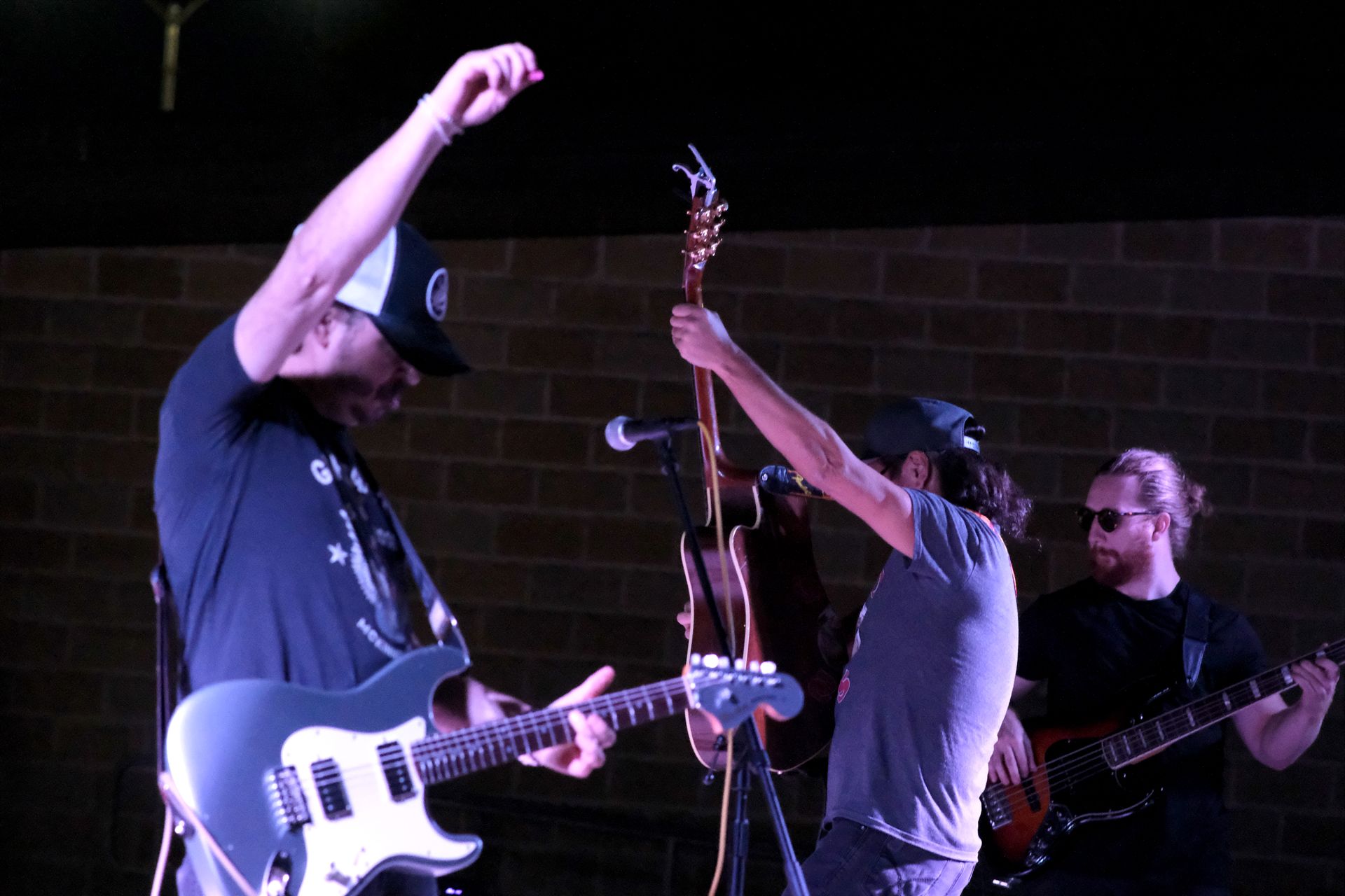 Band on stage, two guitarists with raised arms, one with cap. Bass player in background.