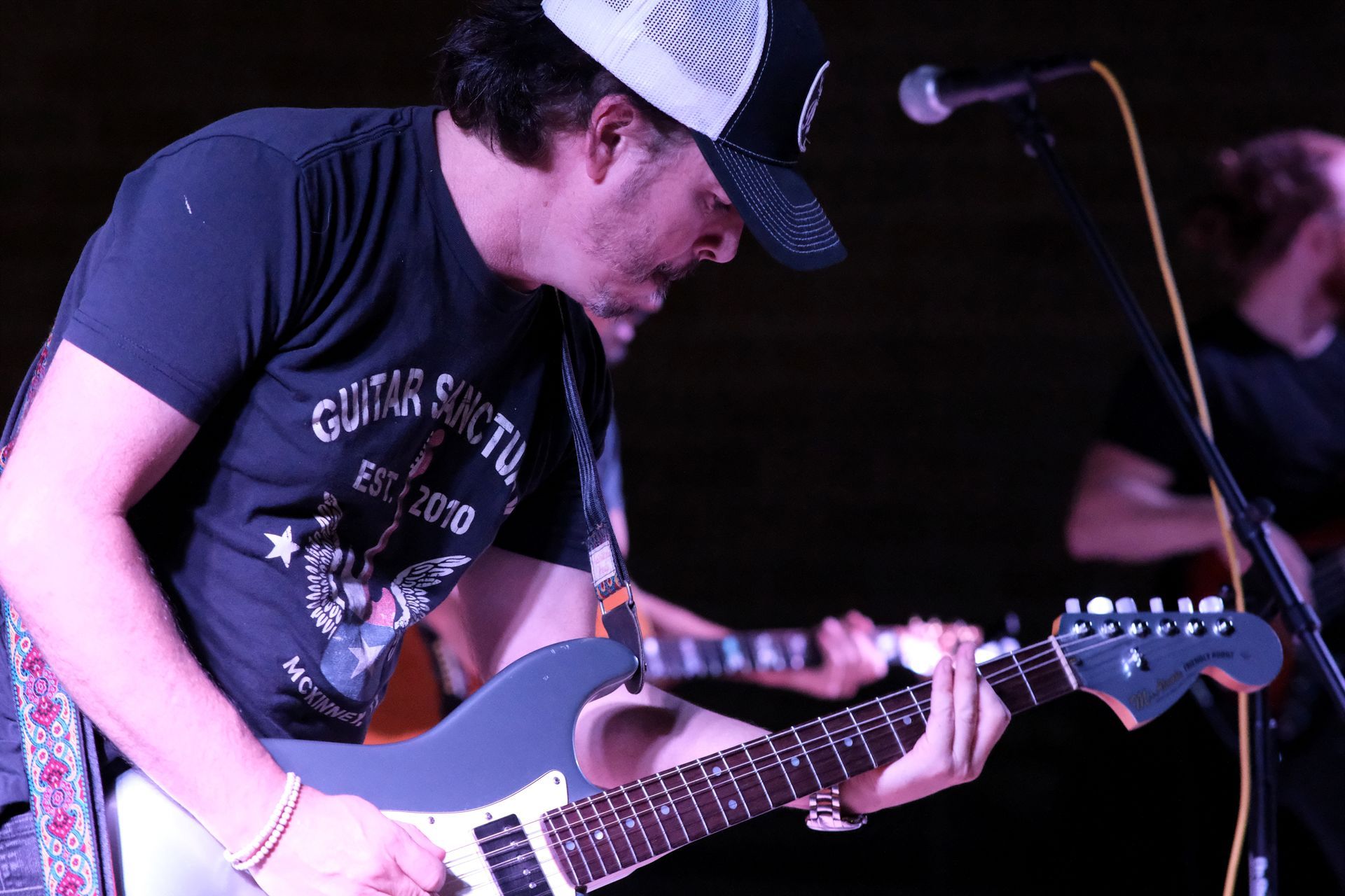 Man playing electric guitar on stage, wearing a hat and t-shirt, lit by stage lights.