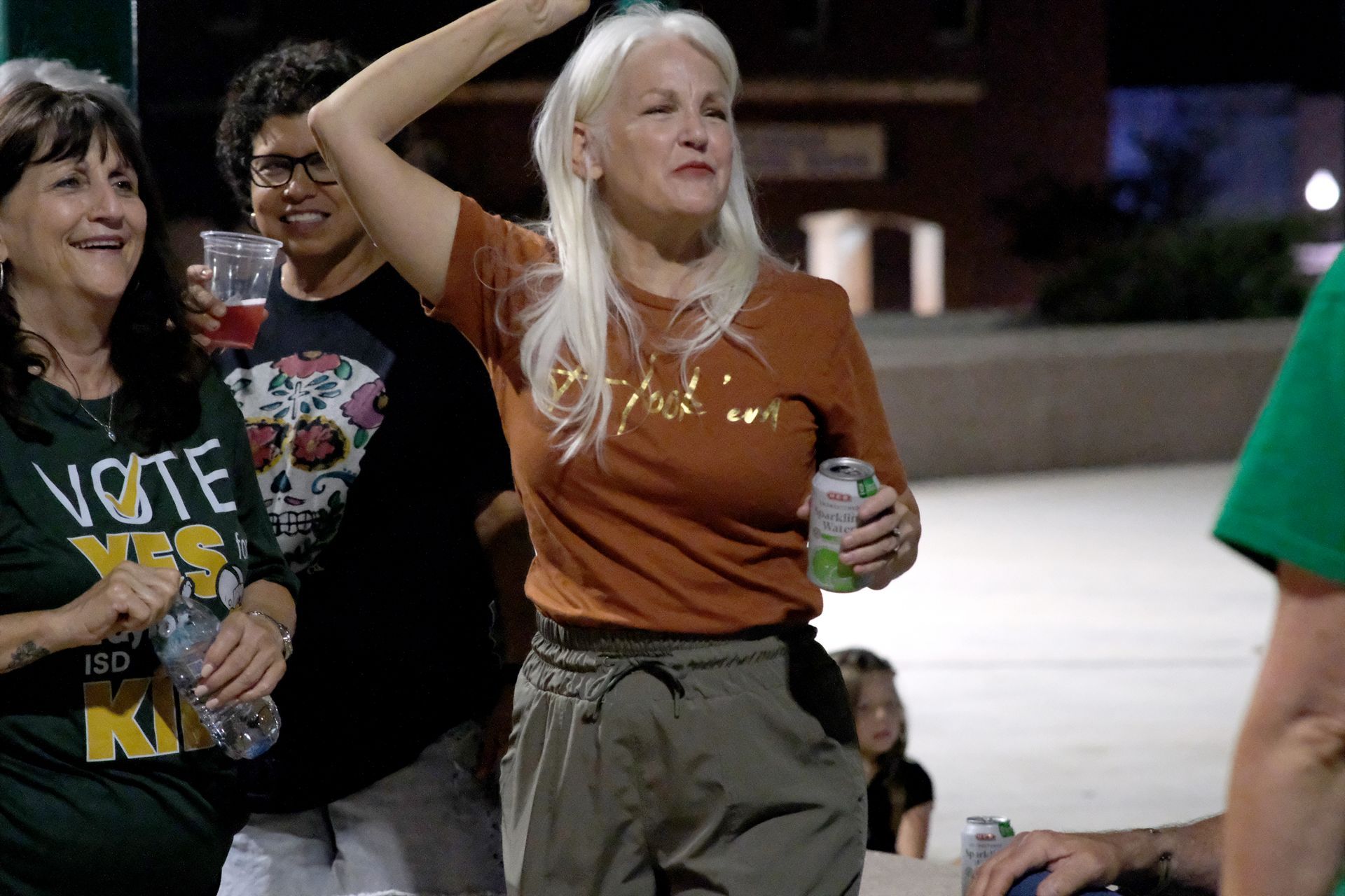 Woman with blonde hair raises arm, celebrating outdoors with others at night.