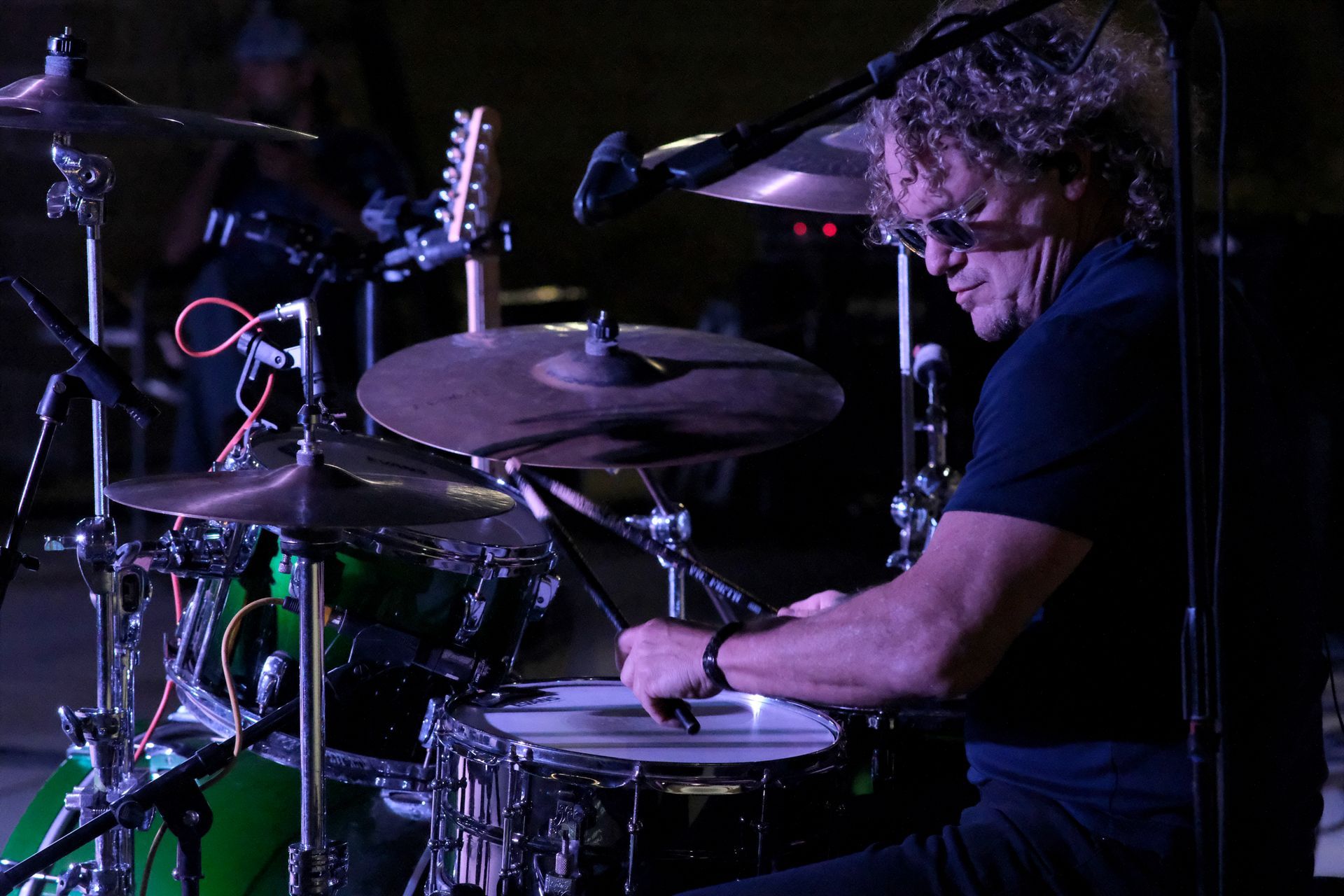 Drummer playing drums on a stage, lit with green and purple lights.