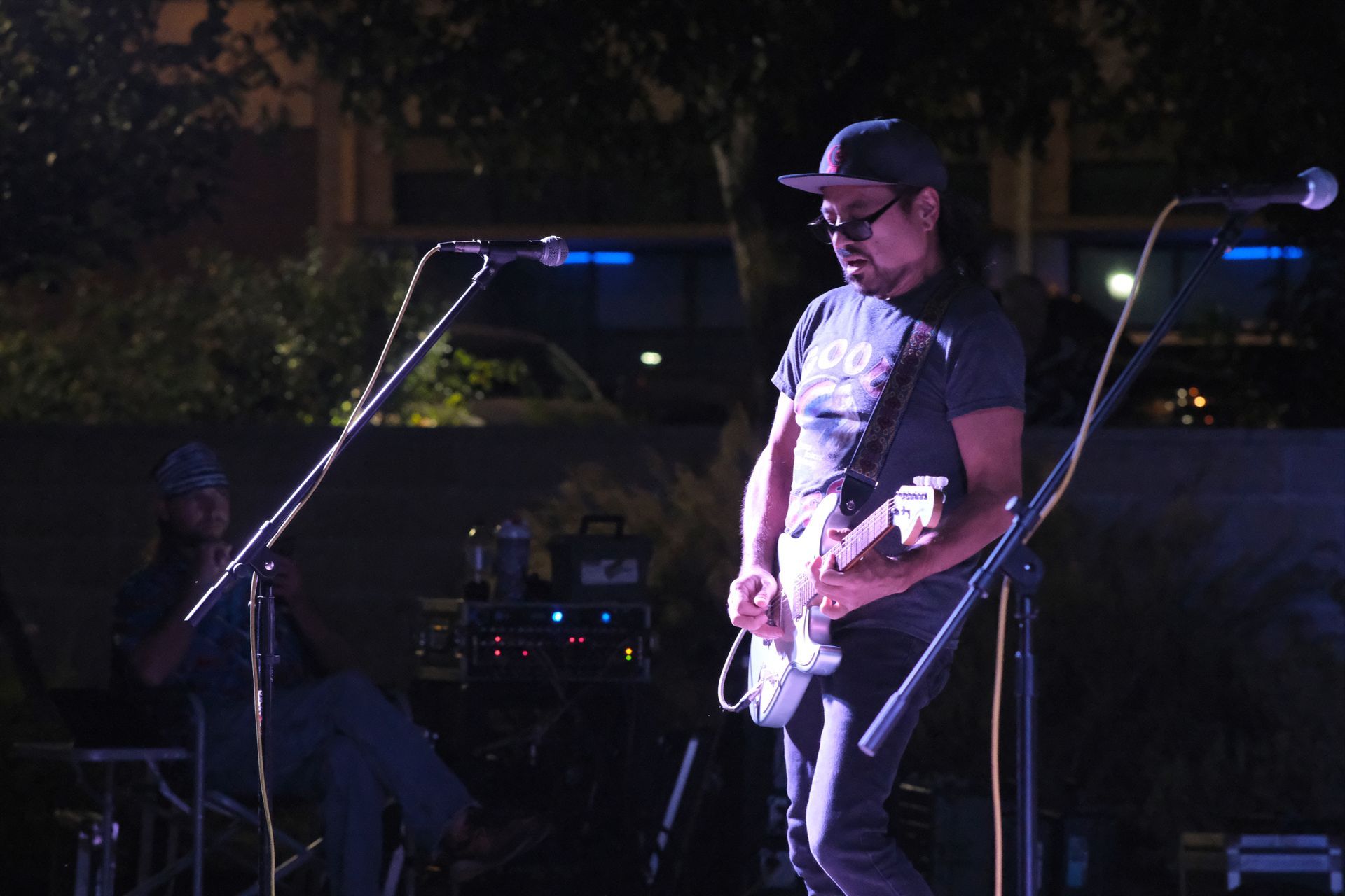 Man playing electric guitar on stage at night, lit by spotlights. Another person visible in background.