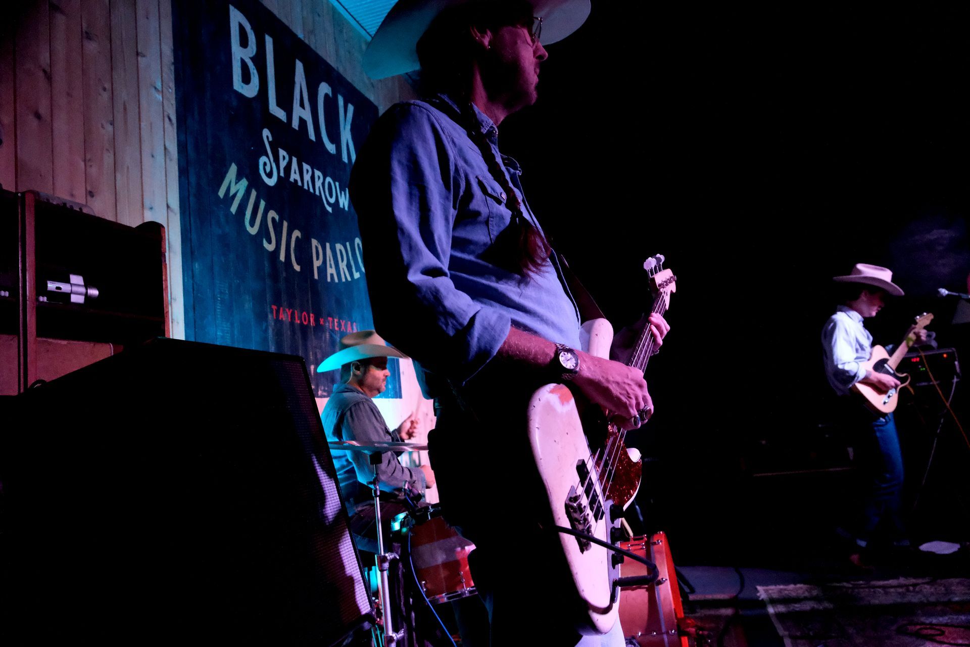 Band performing on stage at the Black Sparrow Music Parlor; three musicians with instruments, stage lit with blues and pinks.