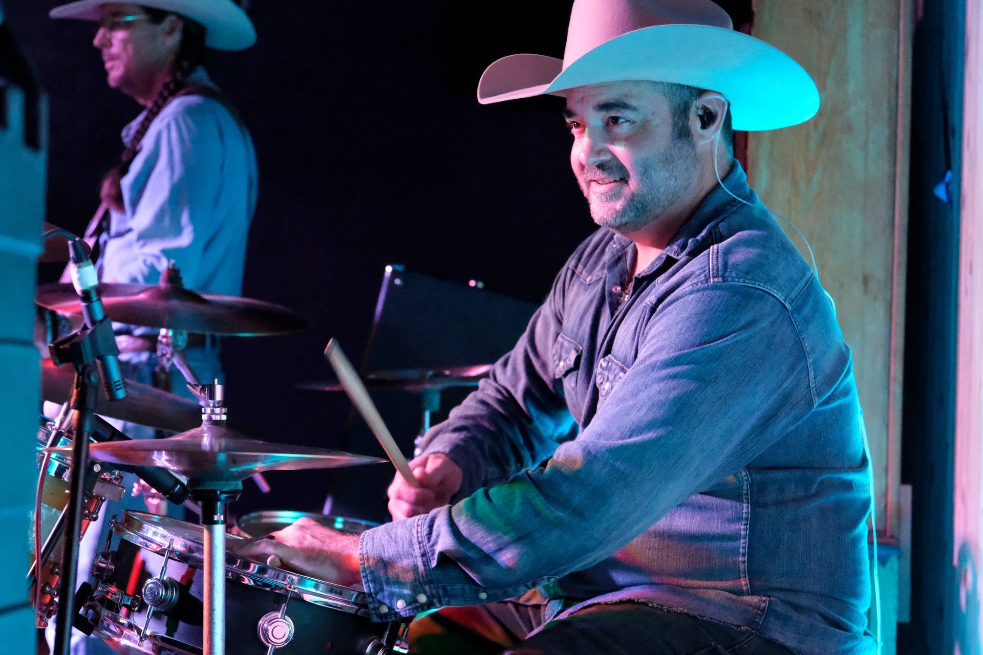 Drummer in a cowboy hat playing drums onstage with a bandmate; blue and purple lighting.