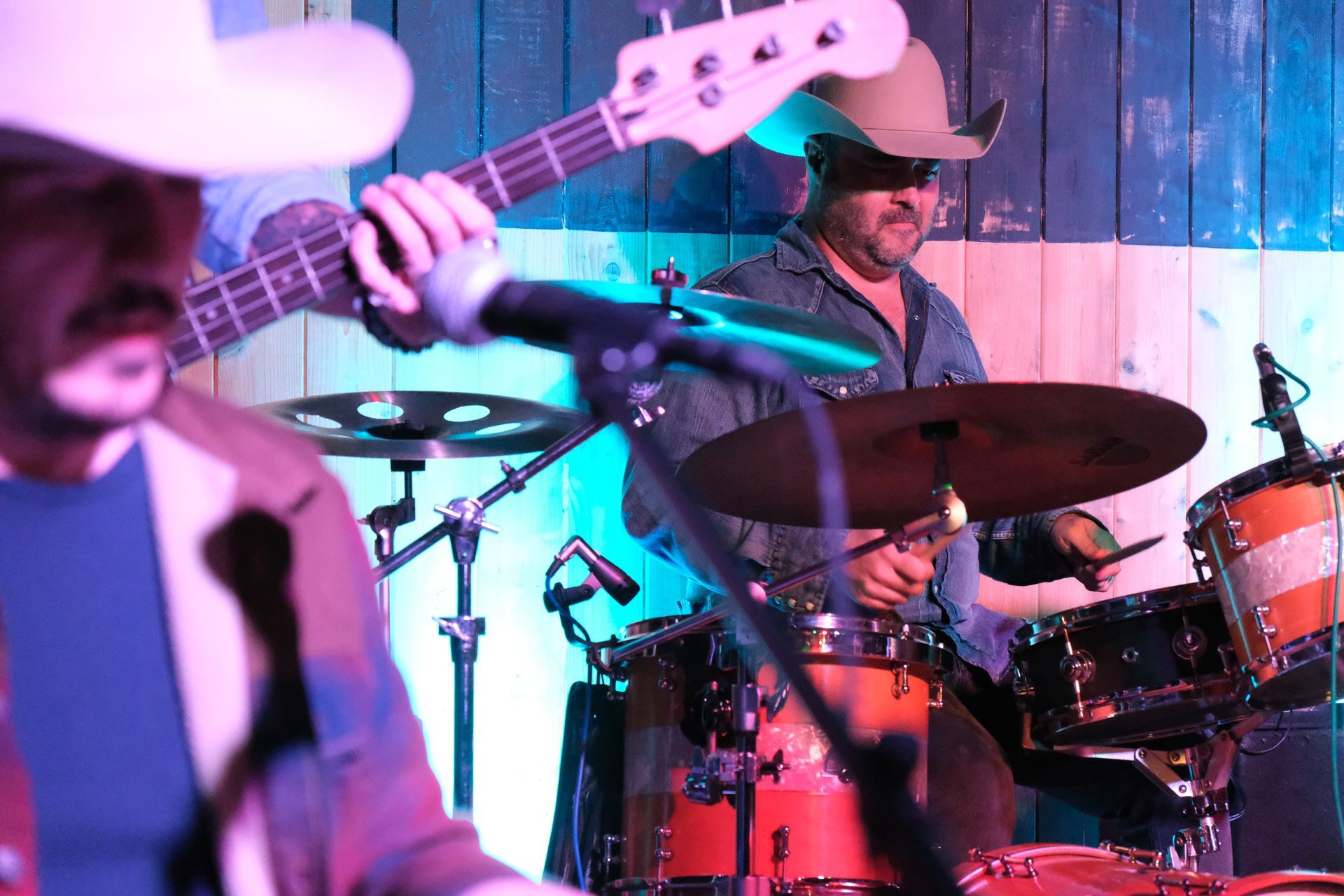 Band playing music on a stage; drummer in a cowboy hat, bassist, and another musician in the foreground.