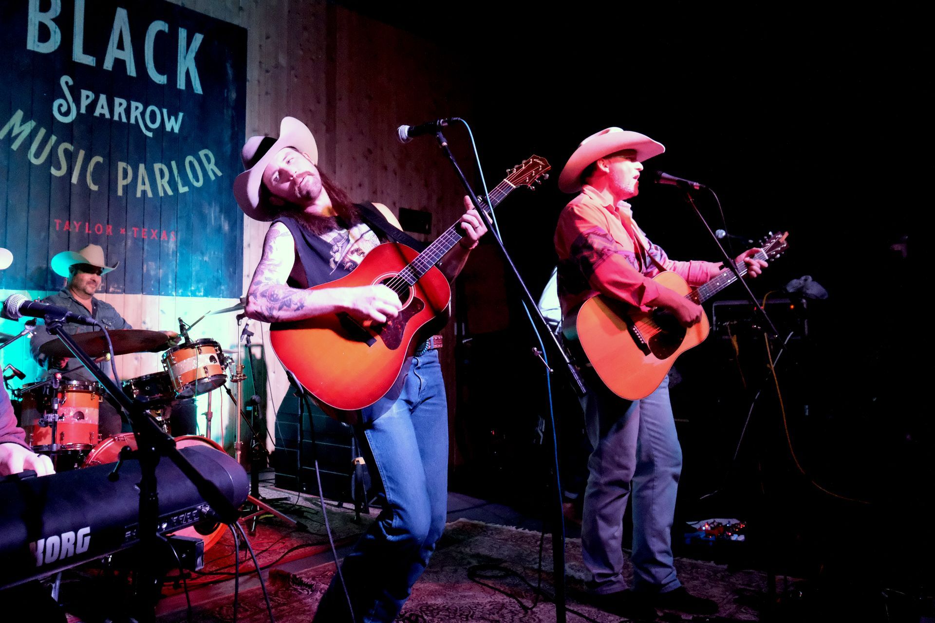 Band performing on stage at Black Sparrow Music Parlor. Two guitarists wearing cowboy hats sing; drummer in background.