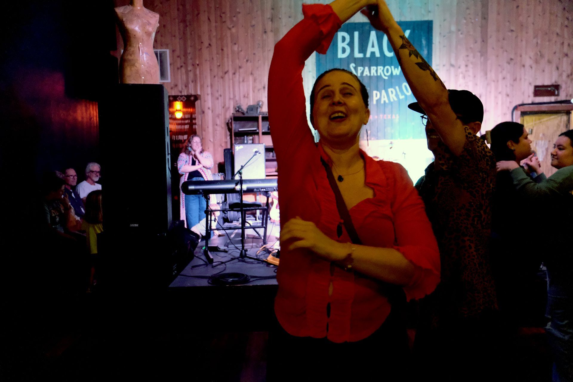 Woman dances joyfully at a live music venue, arm raised. Red shirt, happy expression, stage in background.