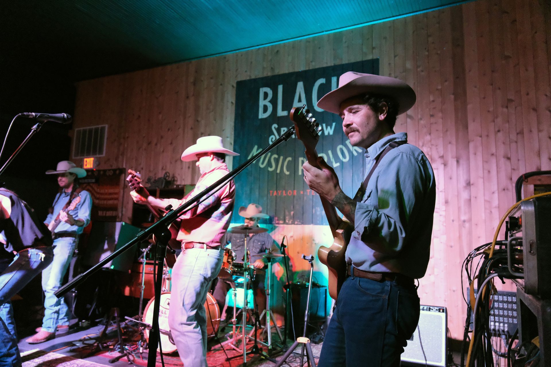 Band playing guitars and drums on stage, all wearing cowboy hats. Wooden backdrop, stage lighting.