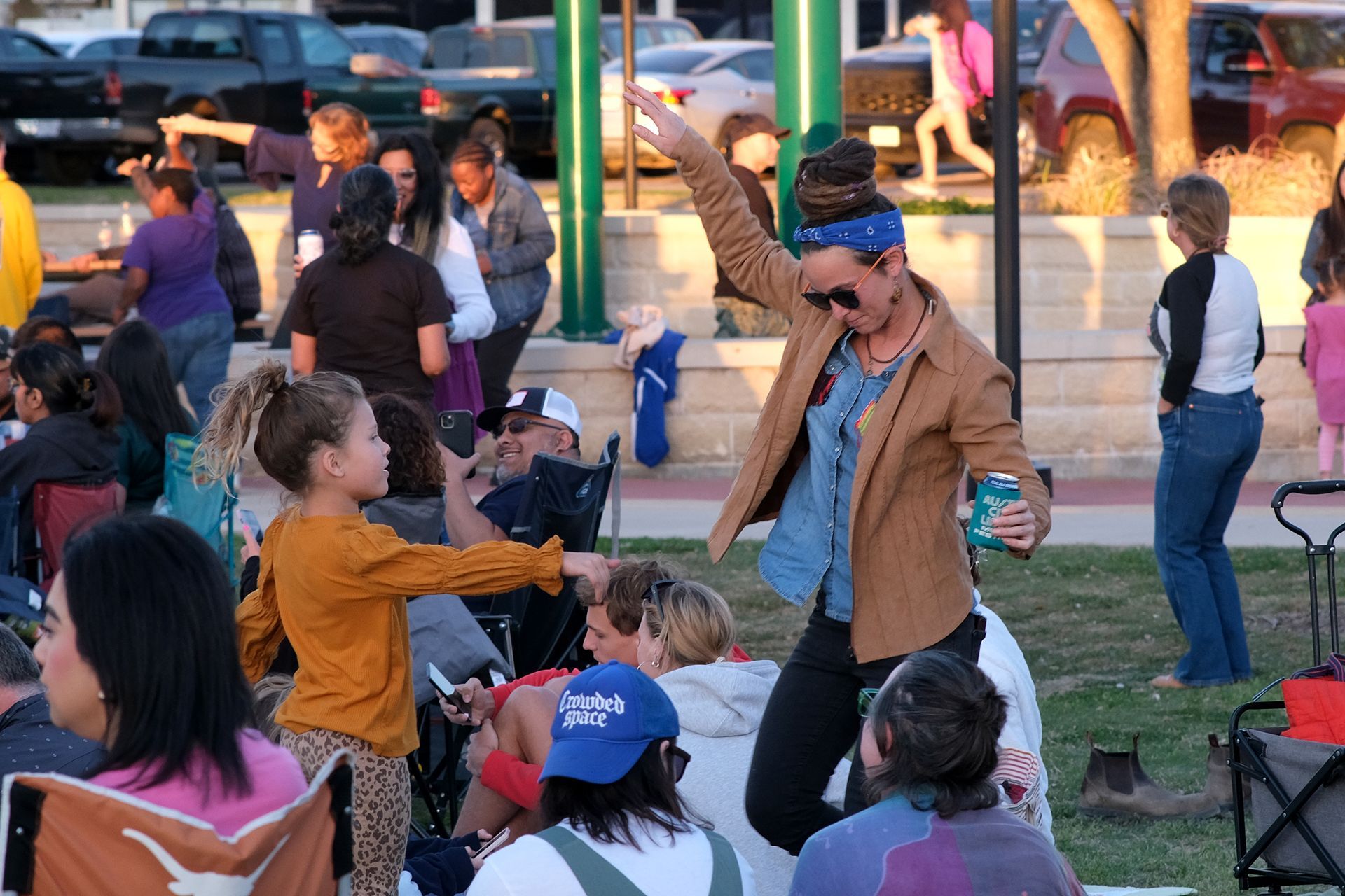 Woman dances, arms raised, with child on grass during outdoor event.
