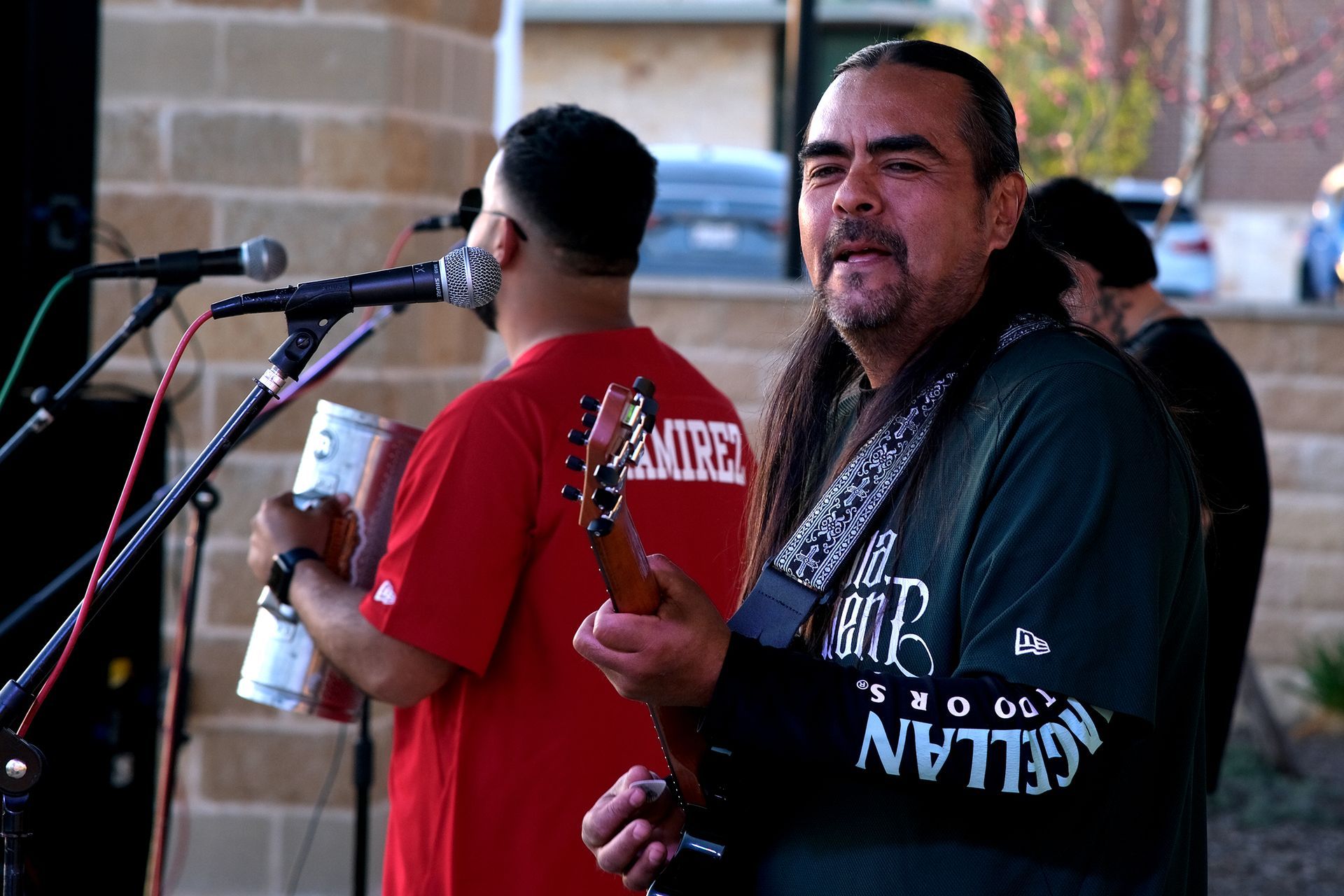 Band performing outdoors; guitarist in green shirt, conga player in red. Microphones, brick building in background.